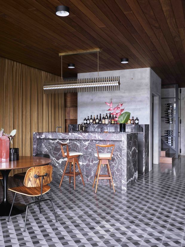 A sophisticated bar and dining area with a dramatic veined stone bar front, timber-lined ceiling and walls, patterned geometric floor tiles, and a sculptural suspended bottle rack. The space includes bar stools, a round dining table with wooden chairs, a drinks display, concrete partition walls, and a glimpse of a stairway and wine storage beyond. The palette is dark, warm, and richly layered with marble-like stone, timber, and monochrome geometric flooring.