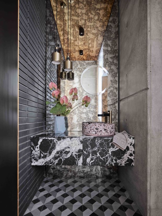 A narrow, dramatically styled powder room with a wall-mounted black-and-white veined stone vanity, pink terrazzo vessel basin, round mirror, black fixtures, pendant lights, patterned floral wallcovering, dark brick-look wall tiles, raw concrete side wall, and bold geometric black, white, and grey floor tiles. A small floral arrangement adds a muted pink and green accent.