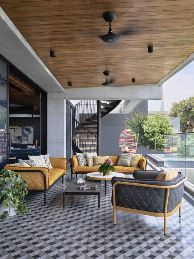 Covered outdoor lounge terrace with mustard and black upholstered seating, a geometric patterned tiled floor, concrete structural beams, a warm timber-lined ceiling with ceiling fans and recessed lights, and a sculptural spiral staircase. Glazing and a glass balustrade open the space to greenery and a pool or lower-level outdoor area. The furnishings and finishes create a bold contemporary setting.