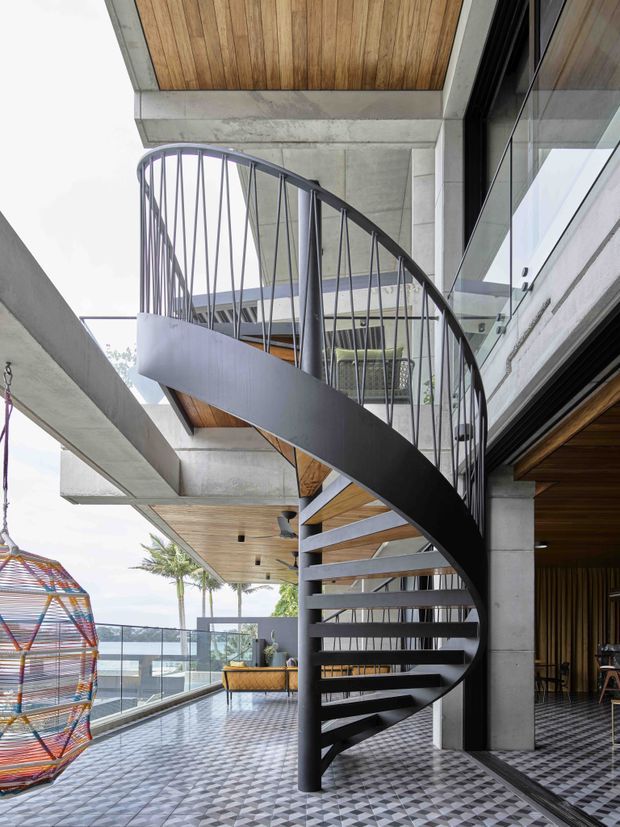 A contemporary multi-level interior featuring a striking black metal spiral staircase with slender vertical balusters and open treads. Exposed concrete columns and beams frame the space, while warm timber-lined ceilings and soffits contrast with the grey structure. Large glazed openings connect to a balcony or terrace overlooking a coastal setting, and patterned grey-and-white floor tiles extend through the room. A colorful woven hanging chair is visible at the left.