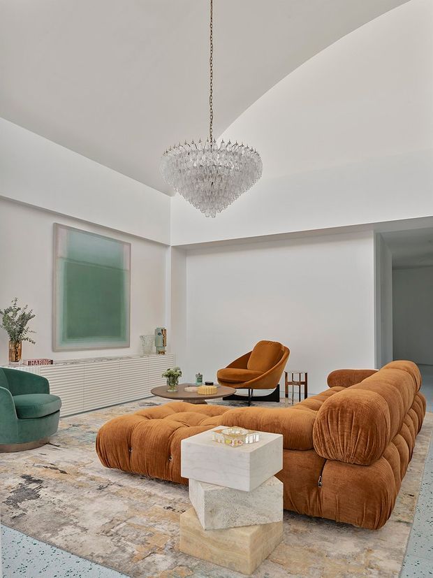A refined contemporary living room with a large sculptural rust-orange modular sectional, matching lounge chair, green accent chair, sculptural stone coffee table, pale patterned rug, ribbed white built-in cabinetry, abstract artwork, and a prominent crystal chandelier. The room has a bright, neutral shell with a high stepped ceiling and a calm yet luxurious atmosphere.