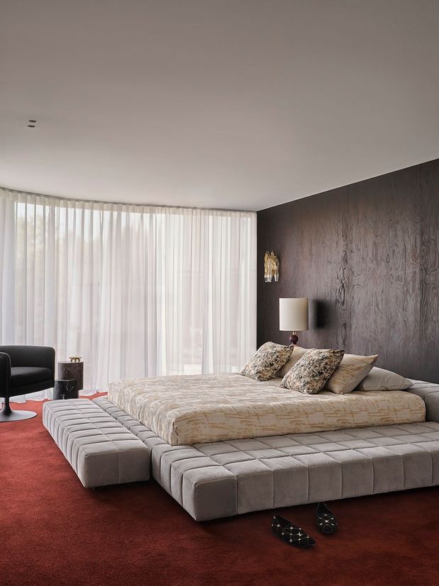 A spacious, refined bedroom featuring a low-profile upholstered platform bed with a matching quilted surround and bench seating. The room has deep red carpet, a dark textured feature wall, sheer full-height curtains across a curved or broad glazed window, a sculptural black lounge chair, and warm bedside lighting. The palette and plush materials create a luxurious, dramatic atmosphere with contemporary styling.