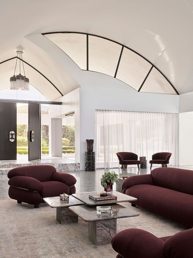 A bright, spacious formal living room or foyer with a dramatic curved vaulted ceiling featuring dark metal framing and translucent panels. The room has white walls, full-height sheer curtains, expansive glazing, polished stone or terrazzo flooring, a marble-clad raised entry platform, and sculptural dark timber or black doors. Deep burgundy upholstered lounge chairs and sofas surround layered marble coffee tables, creating a luxurious, refined interior.