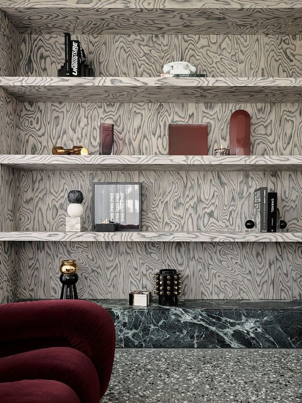 A highly styled interior vignette featuring full-height and layered built-in shelving clad in dramatically figured grey timber veneer. The shelves display books, sculptural objects, framed artwork, and decorative accessories. A dark green-black veined stone ledge and speckled terrazzo floor contrast with a deep burgundy curved lounge chair in the foreground. The composition has a luxurious, moody, contemporary feel with strong graphic material patterns.
