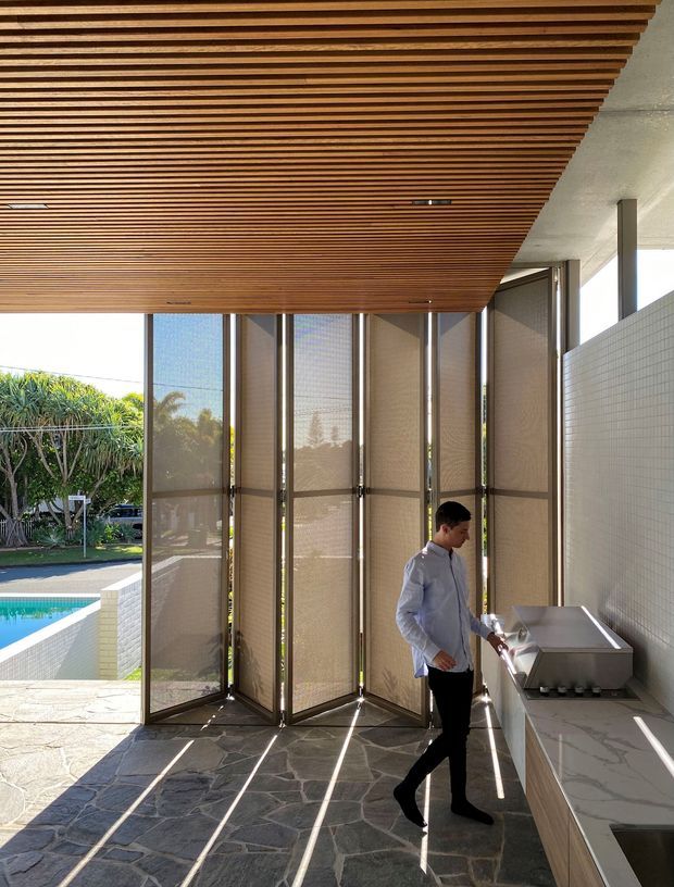 A covered outdoor entertaining area beside a swimming pool, featuring a built-in barbecue and pale stone benchtop, a slatted timber ceiling, dark irregular stone paving, tiled walls, and folding screened glass doors opening to a landscaped garden. The space has strong indoor-outdoor connections and warm natural materials.