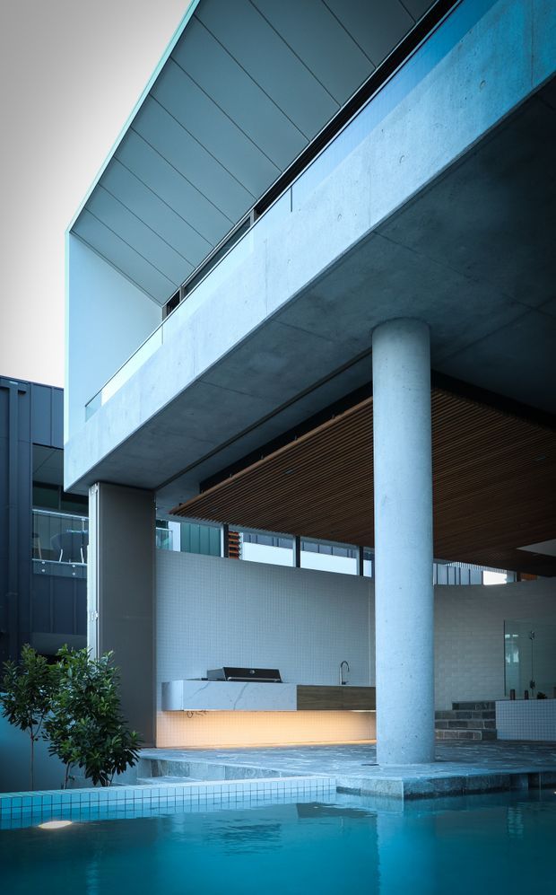 A modern multi-level outdoor entertaining area centred around a swimming pool, with a concrete cantilevered structure supported by a large round column. The space features a timber slatted ceiling, pale tiled walls, glass balustrades, an integrated outdoor kitchen or bar, and steps leading toward the pool. The image has a cool blue-toned, contemporary architectural aesthetic.