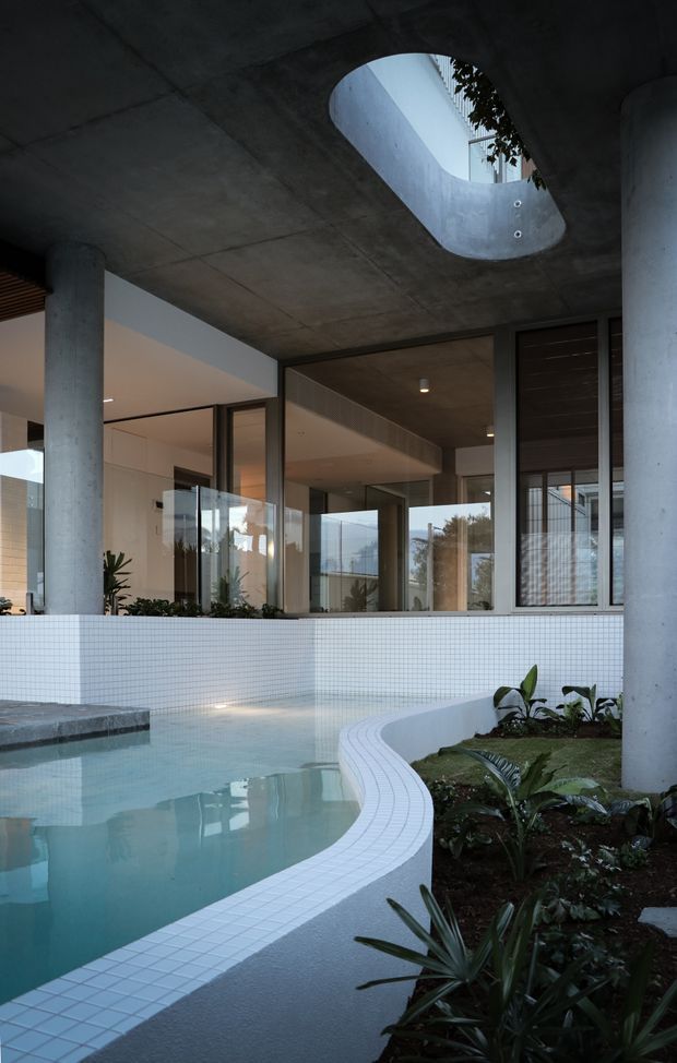 A partially enclosed indoor-outdoor pool courtyard beneath a raw concrete canopy. The space features a curved white small-format tiled pool edge and walls, pale blue water, cylindrical concrete columns, expansive glazed doors and windows, an oval skylight, and integrated planted garden beds. A modern interior is visible beyond the glazing, while the pool and architectural structure dominate the view.