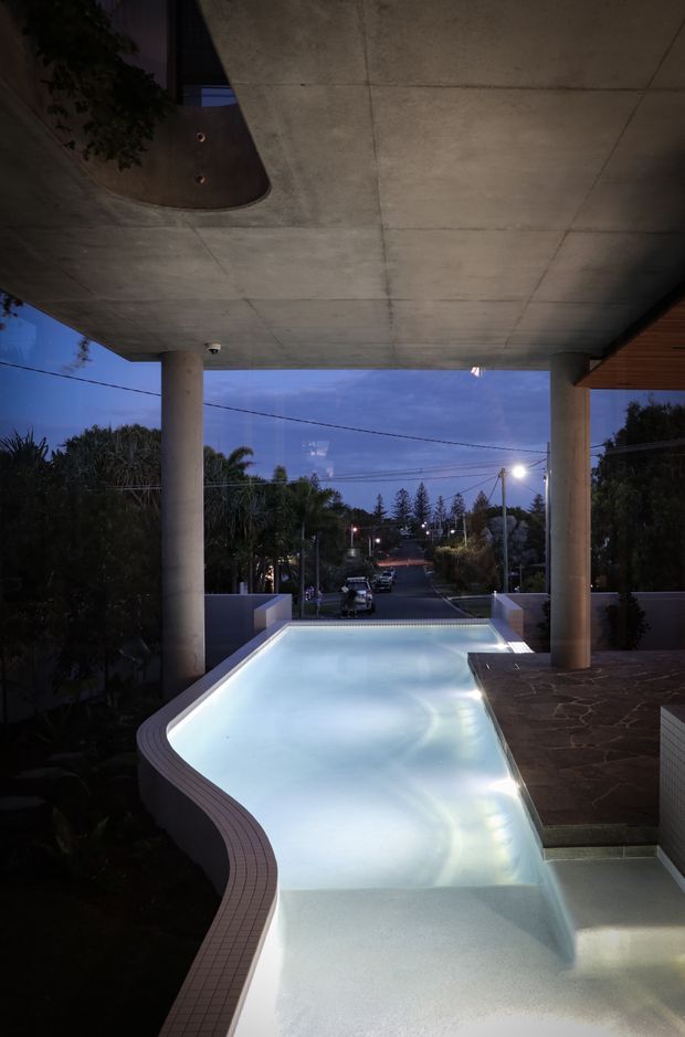 Twilight view from beneath a large concrete-covered outdoor structure, looking over a curving illuminated swimming pool toward a suburban street. The pool has pale tiled coping and integrated steps, with cylindrical concrete columns, a dark stone-paved terrace, timber soffit detailing, and landscaped planting around the edges.