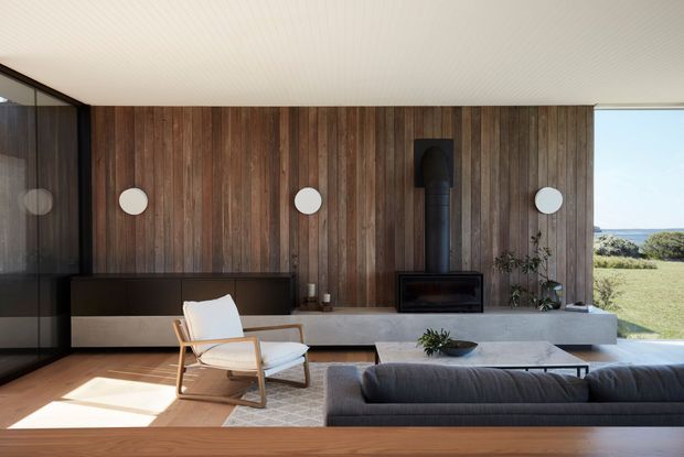 A refined contemporary living room with a full-height dark timber feature wall, integrated low cabinetry, fireplace and round wall lights. A low grey sofa and timber-framed lounge chair face a central coffee table, while large glazing opens to a grassy coastal landscape and ocean view. The room has a low, clean-lined profile and abundant natural light.