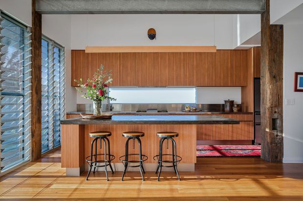 A warm, mid-century-inspired kitchen with a large timber island, three black metal bar stools, extensive vertical timber cabinetry, and a dark stone benchtop. The space features polished timber flooring, exposed timber structural posts, louvred glazing on the left, and a long linear pendant light. A glimpse of a living area with a patterned rug is visible beyond the kitchen.