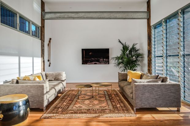 Bright, spacious living room with two facing grey sofas, patterned area rug, timber flooring, wall-mounted television, indoor palm, exposed dark timber posts, and a prominent horizontal concrete or stone beam. Large louvred windows on both sides provide abundant daylight and a strong connection to the exterior. The room combines a neutral contemporary shell with warm timber, woven textiles, and subtle mustard accents.