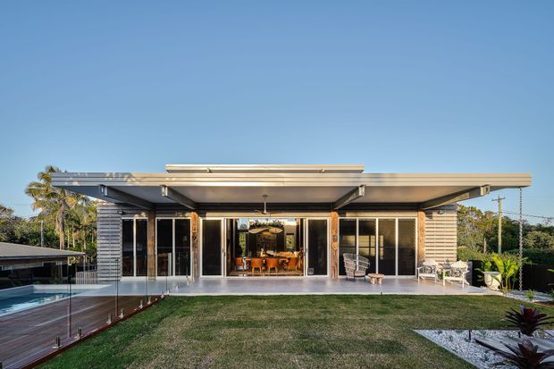Exterior rear view of a low, single-storey modern home with broad flat roof overhangs, extensive floor-to-ceiling glazing and sliding doors, horizontal weatherboard cladding, and a covered outdoor entertaining area. A lawn, pool edge, glass pool fencing, and landscaped garden are visible in the foreground and around the house.