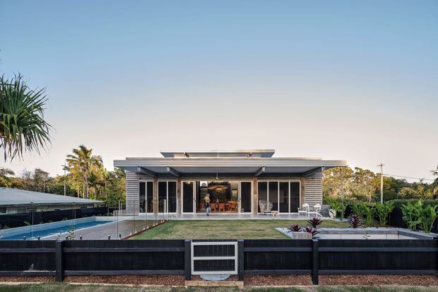 Front exterior view of a single-storey contemporary residence with a broad low-pitched roof, extensive floor-to-ceiling glazing, horizontal cladding, a raised deck, landscaped front yard, swimming pool to the side, and a dark perimeter fence.