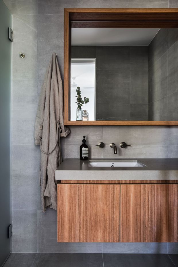 A refined contemporary bathroom vanity with a wall-mounted timber vanity, pale stone benchtop and integrated rectangular basin. Large grey-format wall and floor tiles create a cool, monolithic backdrop, while the timber-framed mirror and warm timber cabinetry add contrast. Dark metal tapware and understated accessories complete the scheme.