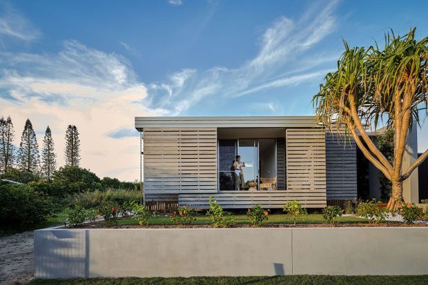 A compact modern single-storey dwelling viewed from the landscaped front garden. The building has a simple flat roof, extensive horizontal timber-like slatted cladding, dark window and door frames, and a recessed glazed central opening. A raised concrete retaining or planter wall runs across the foreground, with tropical and landscaped planting around the home.