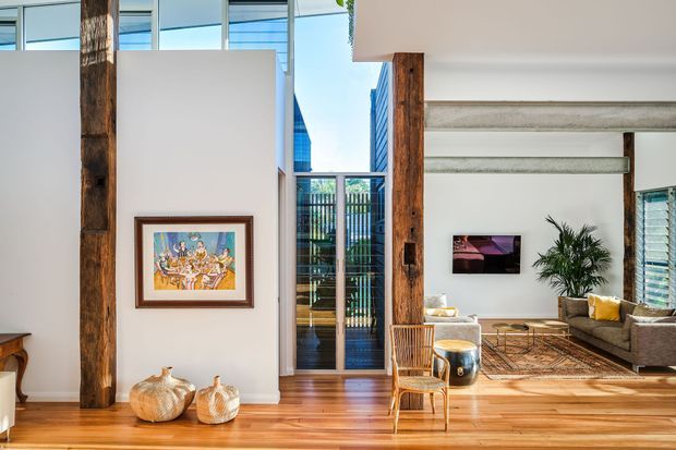 Bright open-plan interior featuring polished timber flooring, tall white walls, exposed rustic timber posts, clerestory glazing, and a double-height central entry or circulation space. A living room with a sectional sofa, armchair, coffee table, rug, and indoor plant is visible to the right, with a dining area partially visible at left. Contemporary furnishings combine with exposed structural timber and abundant natural light.
