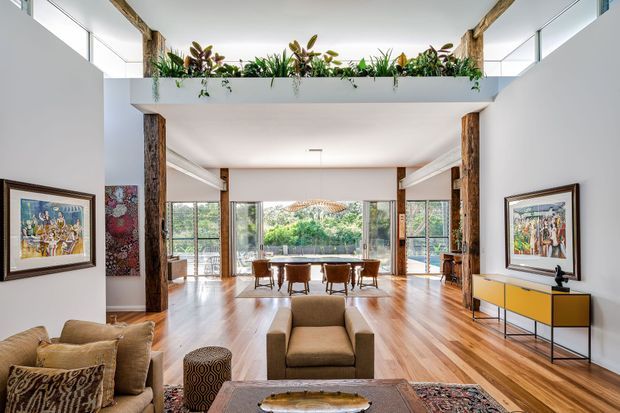 A bright, open-plan interior showing a living room in the foreground flowing into a dining area beyond. The space has polished timber flooring, white walls, large glazed openings, exposed natural timber posts and beams, a double-height void with a planted mezzanine edge, framed artwork, a neutral sofa and armchair, and a colourful low sideboard. The room has abundant natural light and a warm, refined contemporary character.