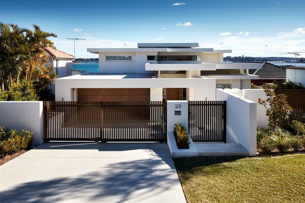 Street-facing view of a contemporary multi-level residence with crisp white rendered walls, strong horizontal roof and balcony lines, dark vertical metal screening, and a timber-toned garage door. A gated driveway and landscaped front garden lead to the entrance. Palm trees, blue sky, and glimpses of neighboring homes and water or distant landscape are visible in the background.