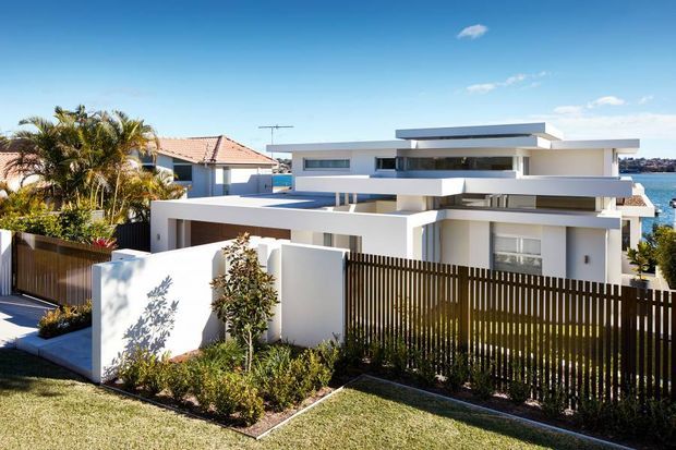 Exterior view of a contemporary multi-level coastal residence with crisp white rendered walls, broad flat roof planes, projecting balconies, and extensive horizontal glazing. A timber slat boundary fence and landscaped garden occupy the foreground, with glimpses of water and neighbouring homes beyond.
