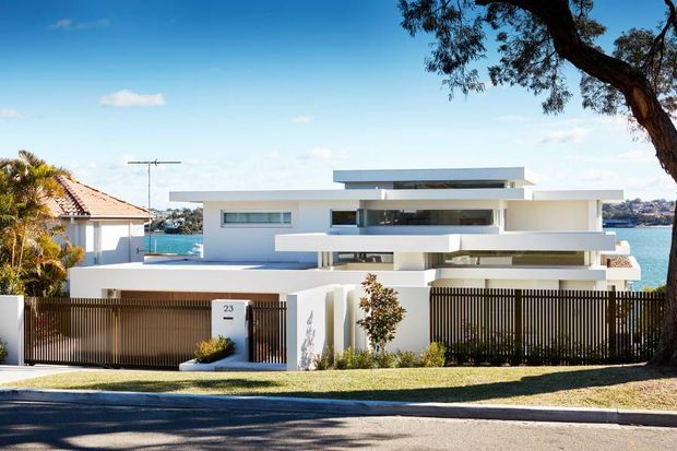 Street-facing exterior view of a large contemporary waterfront residence with crisp white horizontal forms, broad glazing, layered flat rooflines, balconies, and a tall dark vertical-slat boundary fence. A neighbouring tiled-roof house and water are visible behind the property.