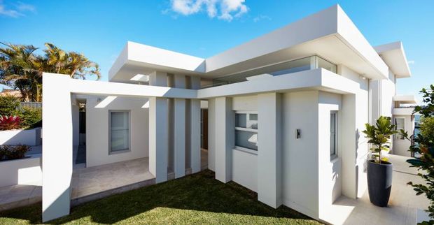 Exterior view of a contemporary two-storey residence with crisp white rendered walls, strong horizontal and cubic forms, deep roof overhangs, covered outdoor circulation, and large glazed openings. A small landscaped lawn and potted tropical planting sit around the house under a bright blue sky.