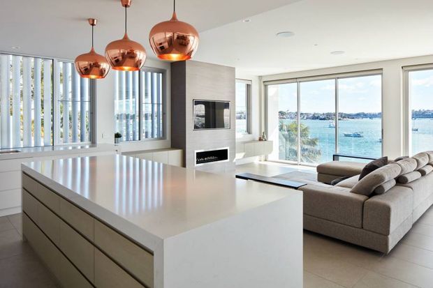 Bright contemporary open-plan kitchen and living room overlooking a waterfront. A large white island with integrated cabinetry dominates the foreground, complemented by copper pendant lights, pale flooring, a minimalist fireplace and television wall, and a large neutral sectional sofa. Floor-to-ceiling glazing provides expansive water views.