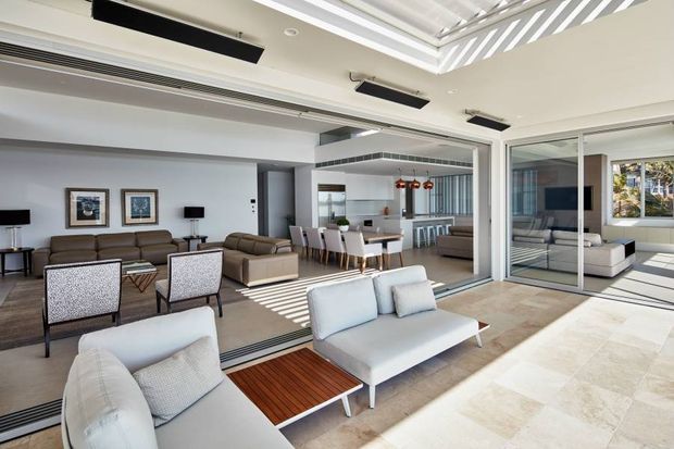 Bright contemporary open-plan living area with a connected dining space and kitchen, viewed through wide glazed sliding doors. The room features pale upholstered seating, a neutral rug and floor, dark timber accents, framed artwork, linear black ceiling fixtures, and strong striped sunlight from a roof opening or louvred shading. The kitchen includes a central island with bar seating and white cabinetry.