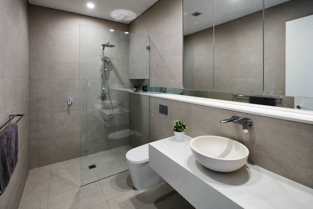 Contemporary bathroom featuring large-format beige-grey tiled walls and floor, a frameless glass walk-in shower, wall-hung toilet, floating white vanity with a round vessel basin, wall-mounted chrome tapware, and a large mirrored wall. Recessed ceiling lighting creates a clean, refined appearance.