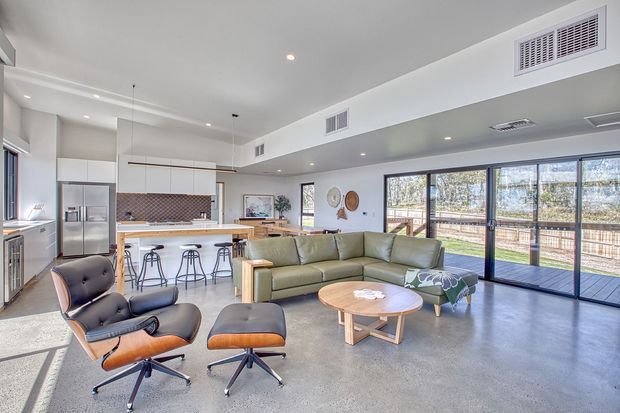 A bright, open-plan contemporary living area with a connected kitchen and dining zone. The space features polished concrete flooring, white cabinetry, a tiled kitchen splashback, extensive black-framed glazing and sliding doors opening to an outdoor deck, plus mid-century-inspired lounge furniture and a timber coffee table.