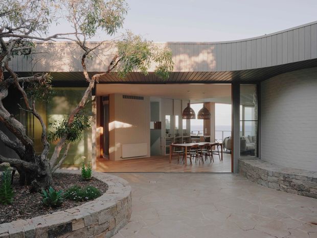 Exterior view of a low-slung contemporary house opening onto a paved courtyard. The building has dark vertical metal cladding, a broad curved roofline, large glazed openings, warm timber soffits, and a pale stone-clad wall. Through the open glazing, a dining area with a timber table, chairs, and pendant lights is visible. Mature planting and a curved stone garden wall frame the entrance.