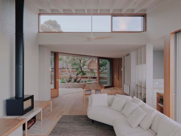Bright open-plan living and dining space with a large white sectional sofa, black freestanding wood heater, light timber flooring, white-painted brick and plaster walls, exposed white ceiling beams, and extensive timber-framed glazing overlooking a landscaped courtyard. Built-in timber joinery and vertical shelving line the right side of the room.