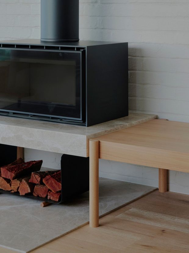 Close-up interior detail of a contemporary wood-burning fireplace with a black metal surround and flue, set into a pale stone or concrete hearth. A white-painted brick wall forms the backdrop, with stacked firewood stored beneath the hearth and a simple light timber console or bench extending to the right. The space has a restrained, material-focused aesthetic with pale timber flooring and neutral finishes.