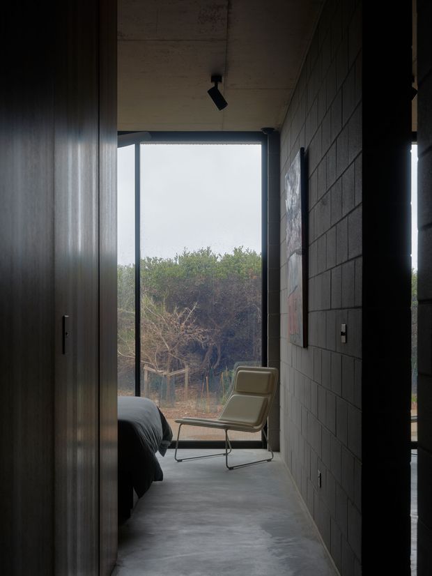 A narrow, moody bedroom or sleeping space with a visible bed edge, dark timber wall or built-in joinery on the left, exposed concrete ceiling and polished concrete floor. A dark concrete-block wall lines the right side, while full-height glazing opens to dense vegetation outside. A minimalist lounge chair sits beside the window, and a small track light illuminates the ceiling.