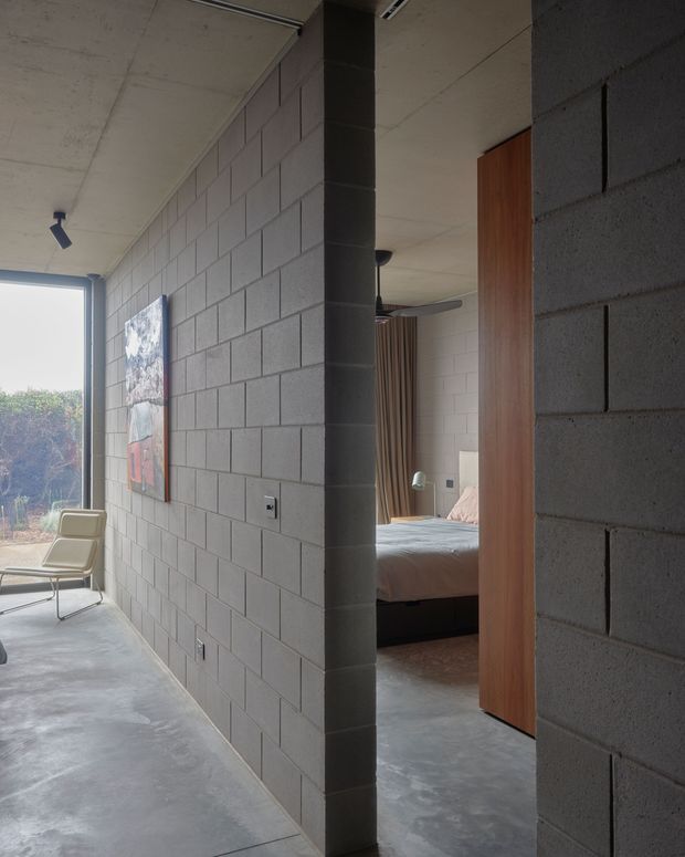 A minimalist interior corridor with exposed grey concrete block walls, polished concrete flooring, and a smooth concrete ceiling. A large glazed opening brings in natural light and views of vegetation, while a lounge chair and artwork sit along the corridor. Through an open doorway, a bedroom with a bed, curtains, and timber-toned door or cabinetry is visible. The space has an industrial, raw-material character with clean modern detailing.
