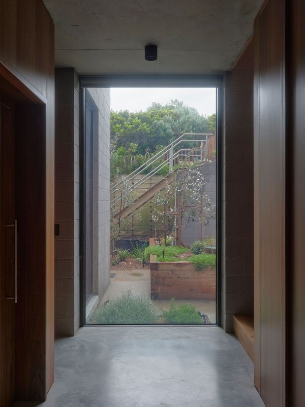 A narrow, minimalist interior passage or entry area framed by dark timber built-in cabinetry and shelving, with a smooth concrete floor and exposed concrete ceiling. A full-height glazed opening at the end provides a strong visual connection to a landscaped courtyard, raised garden beds, and an exterior stairway. The space has a restrained, material-focused architectural character with muted natural tones.