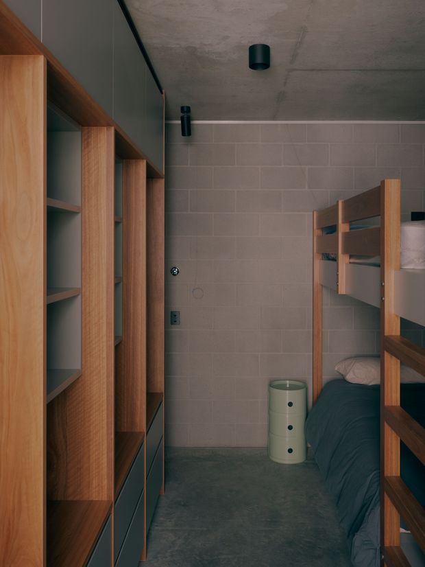 Compact bunk bedroom with a built-in timber storage wall on the left and a timber bunk bed on the right. The room features grey block-pattern wall tiles, a dark concrete-look floor and ceiling, dark teal-grey bedding, and minimalist black cylindrical ceiling and wall lights. The warm timber joinery contrasts with the cool, industrial finishes.