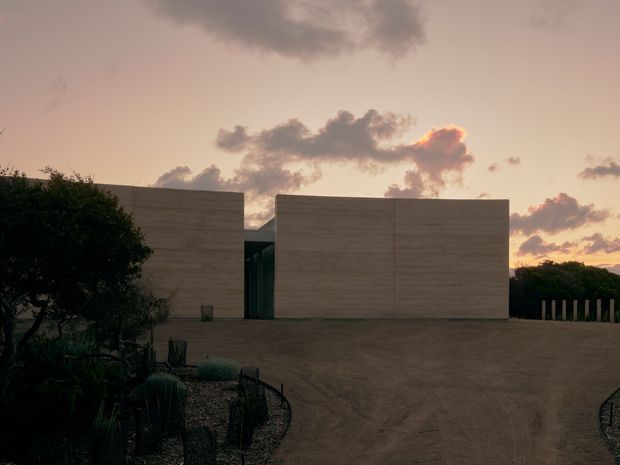 A low, minimalist architectural residence viewed from a broad gravel driveway at sunset. Two large rectangular pale concrete or rendered masonry volumes frame a recessed dark glazed entry, with sparse xeriscape planting and sculptural cacti in the foreground. The flat-roofed composition is stark and monolithic against a warm, cloudy sky.