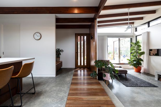 Open-plan contemporary home interior showing a kitchen island with bar stools on the left, a timber-framed entry hall with a glazed front door, and a bright living area beyond. Dark exposed timber beams span the white ceiling, while a timber-plank walkway contrasts with polished concrete flooring. Large glazing, indoor plants, and neutral furnishings create a warm, airy atmosphere.