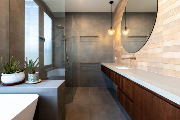 A contemporary bathroom with a walk-in glass shower, freestanding bathtub, long timber vanity with a pale stone benchtop, and a large round mirror. Dark grey wall and floor tiles contrast with a light horizontal tiled feature wall. Pendant lights, black-framed glazing, and potted plants add warmth and detail.