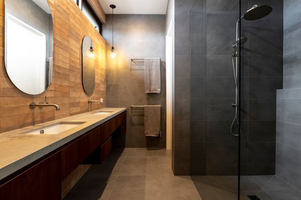 A contemporary bathroom with a long double vanity featuring a pale stone benchtop and dark timber cabinetry, two rounded mirrors, warm wall sconces, and a walk-in shower enclosed by dark grey tiled walls and clear glass. Warm timber wall cladding contrasts with the darker tiled shower and neutral floor tiles.