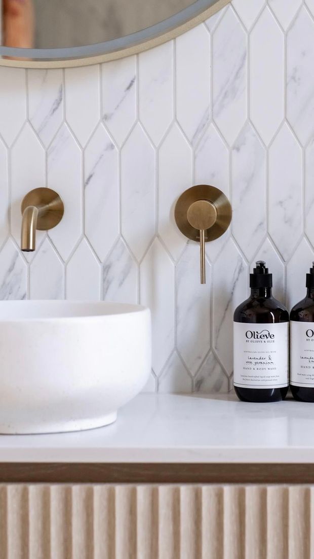 Close-up of a refined bathroom vanity featuring a white vessel basin on a white countertop, wall-mounted brushed brass tapware, and a vertically arranged geometric marble-look tile splashback. A curved mirror with a slim metallic frame is partially visible above, while fluted cabinetry adds texture below.