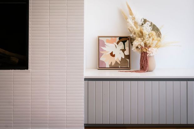 A close-up interior vignette showing a contemporary built-in media cabinet and feature wall. The left side has a wall-mounted black television set against small pale rectangular tiles, while the right side features a white benchtop over vertically panelled grey cabinetry. A framed abstract floral artwork and a vase of dried flowers add warm decorative accents.