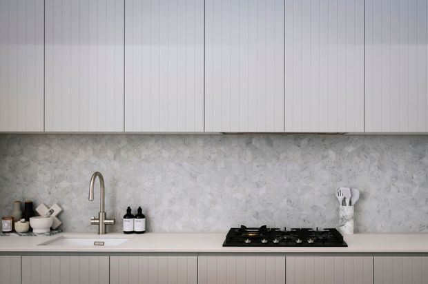 A minimalist contemporary kitchen viewed straight on, featuring vertically fluted light cabinetry above and below a pale stone countertop, a matching pale textured stone splashback, an undermount sink with a brushed metal gooseneck tap, and a black gas cooktop. Neutral tones and clean lines create a refined, understated composition.