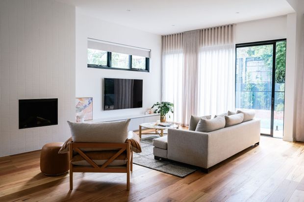 Bright contemporary living room with white vertically panelled walls, a flush black fireplace, wall-mounted television, large upholstered sofa and timber-framed lounge chair. Full-height sheer curtains and sliding glass doors open toward an outdoor area, while warm timber flooring and a simple coffee table create a calm, refined setting.