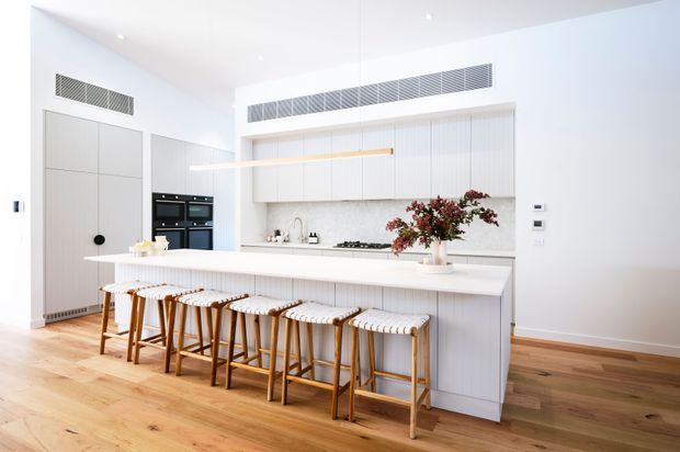 Bright contemporary kitchen with a large white island and extensive seating, full-height white cabinetry, integrated black appliances, a linear pendant light, and light timber flooring. The space has a clean, airy, highly functional design with subtle vertical panelling and warm timber stools.