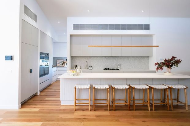 Bright contemporary kitchen with a long white island and integrated seating for five or more, white flat-panel cabinetry, a pale stone-look splashback, timber flooring, and a concealed appliance wall. The space has high ceilings, recessed lighting, linear ventilation, and a clean, airy appearance.