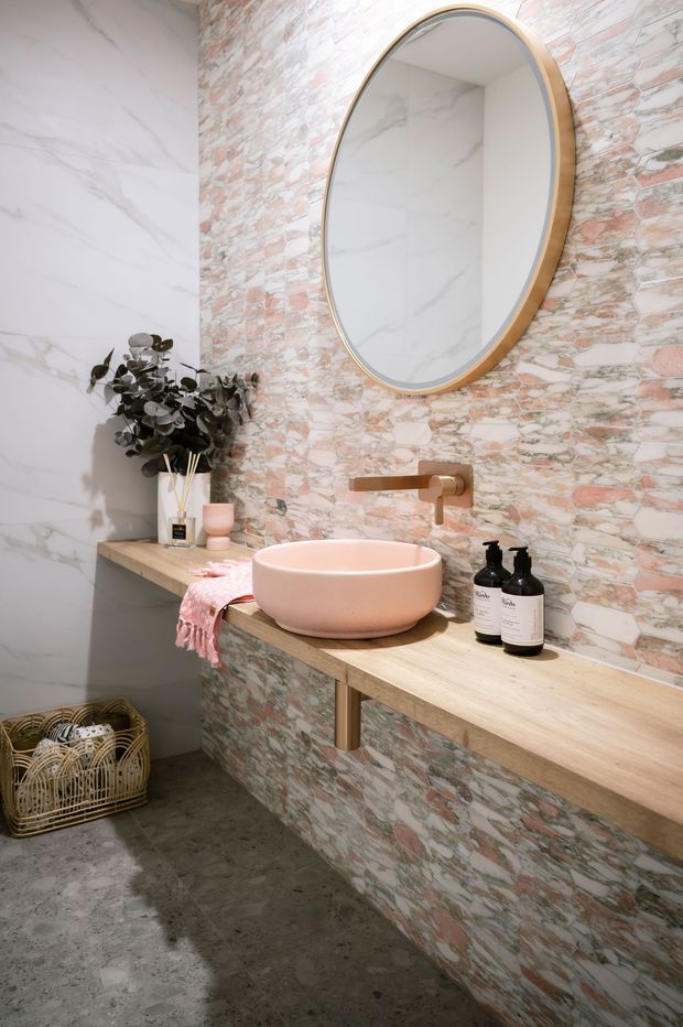 A compact, warmly styled bathroom vanity with a wall-mounted timber benchtop and integrated storage front clad in textured pink-and-white stone mosaic tile. A round vessel basin, brushed brass wall-mounted tapware, and a large oval mirror with a timber-toned frame create a soft contemporary look. Large pale marble-look wall tiles and a polished concrete floor complete the space, with subtle decorative accessories and a woven basket.