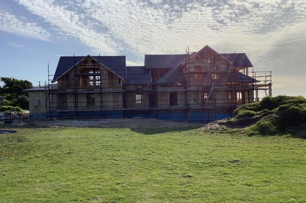 A large multi-gabled house under construction, shown from across a grassy site. The building has a pitched roof, timber-framed gable ends, extensive scaffolding, and partially completed exterior walls and openings.