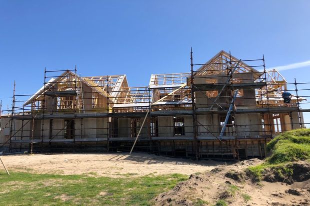 Exterior construction view of a large multi-level timber-framed house under construction, with exposed roof trusses, multiple gabled roof sections, extensive scaffolding, and unfinished wall and window openings. The building sits on a grassy, partly earth-covered site beneath a clear blue sky.