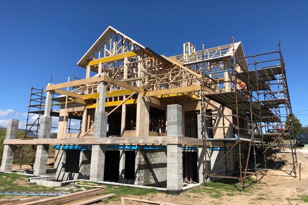 A multi-storey residential building under construction, with exposed concrete block and concrete structural elements, extensive timber roof framing, scaffolding, and unfinished openings. The building has a pitched gable roof and appears to be in an active framing and structural construction phase.